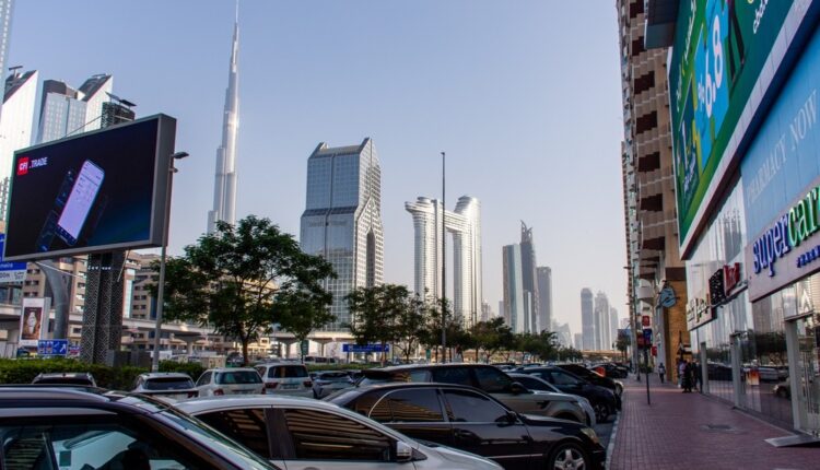 : A view of a bustling street in Dubai with modern skyscrapers, including the Burj Khalifa, visible in the background. There are cars parked along the sidewalk and large digital billboards showcasing advertisements.