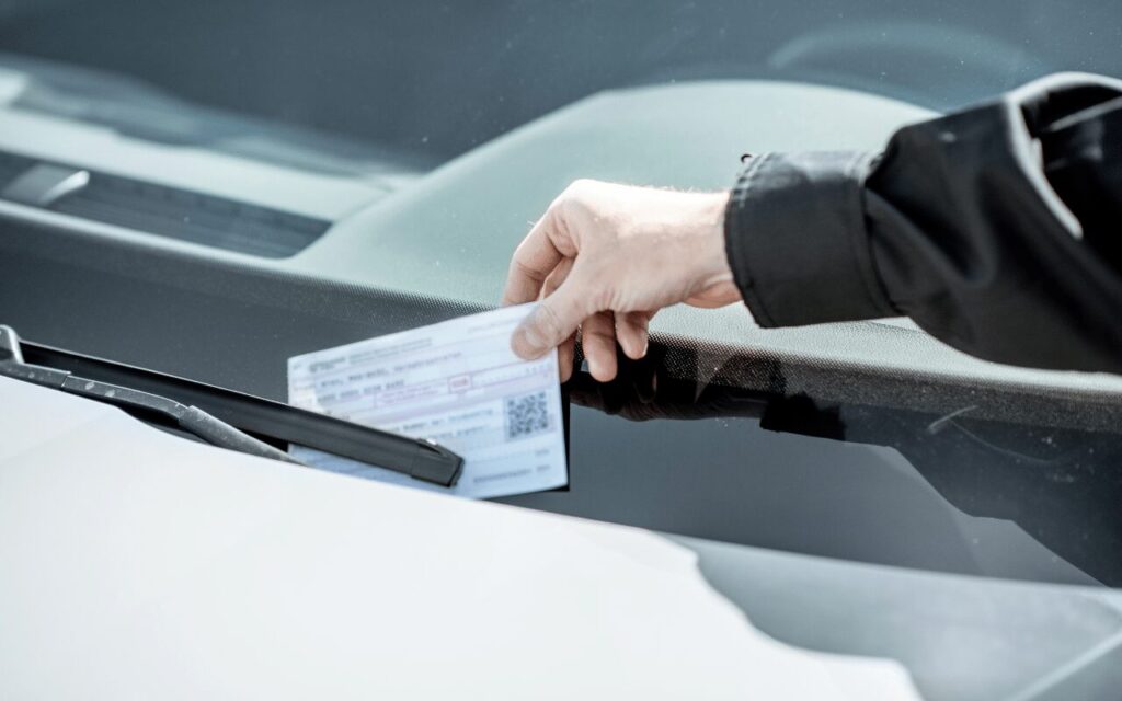 A hand placing a traffic fine ticket under the windshield wiper of a parked car, symbolizing penalties for traffic violations.