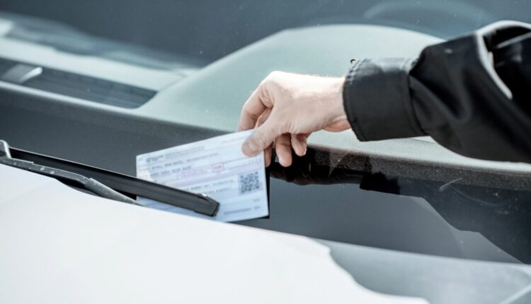 A hand placing a traffic fine ticket under the windshield wiper of a parked car, symbolizing penalties for traffic violations.