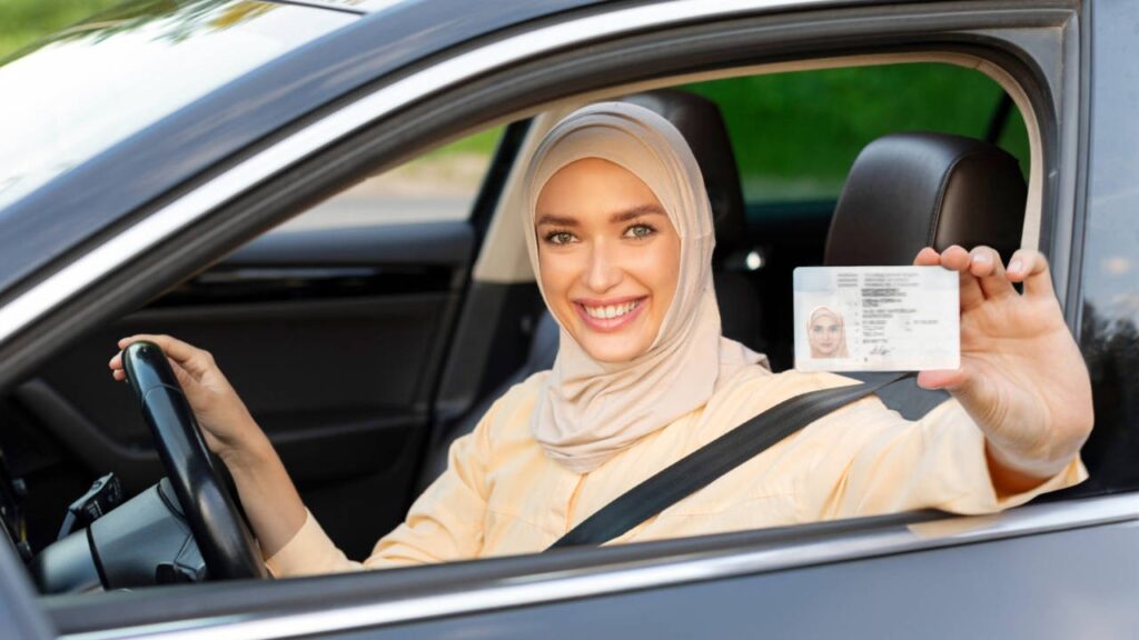 Smiling woman in a hijab sitting in a car holding her driving licence card, representing successful Dubai driving licence management.