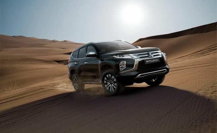 A silver Mitsubishi SUV driving on a desert road surrounded by sand dunes under a bright blue sky, representing strength and reliability in tough environments.