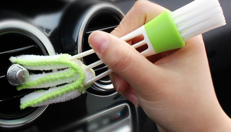 A person using a dual-ended cleaning brush to remove dust from a car air conditioning vent, showing an effective DIY method for car AC maintenance.