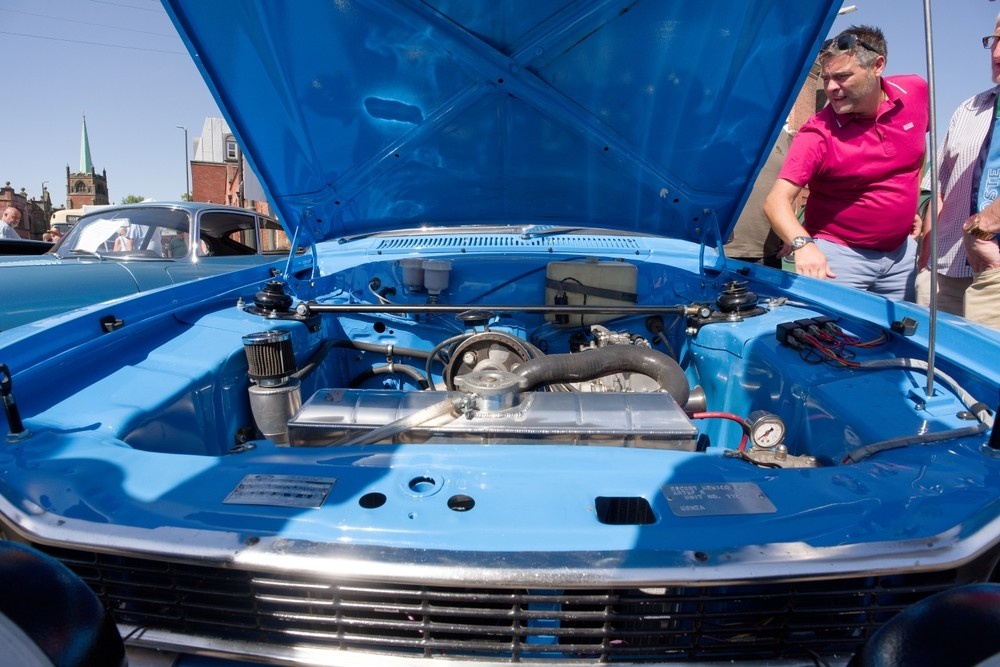 "Checking the engine coolant reservoir under the hood of a classic car without opening the radiator cap.