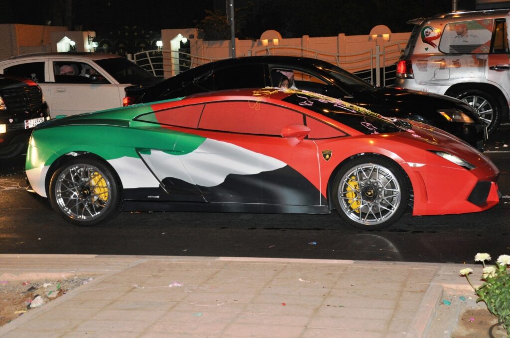 A Lamborghini sports car decorated with a full UAE National Day wrap in red, green, white, and black, parked on a street at night among other decorated vehicles during celebrations.