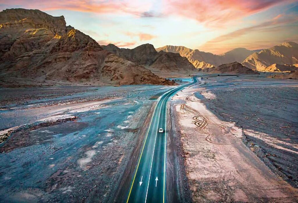 A silver luxury 4x4 SUV parked on a scenic overlook of the winding Jebel Jais mountain road during a golden winter sunset.