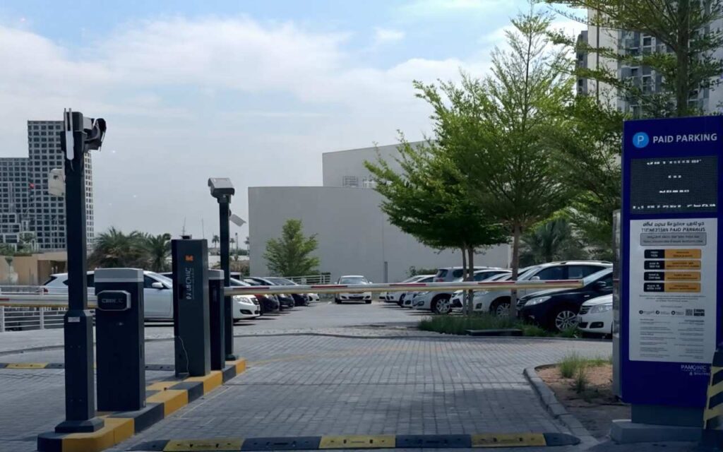 A modern Parkonic automated parking entrance featuring a blue digital signage board, boom gate barriers, and parked cars in a clean urban parking lot.