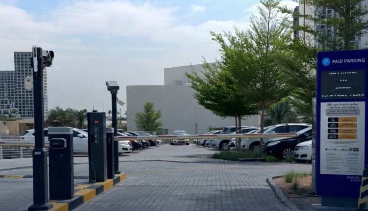 A modern Parkonic automated parking entrance featuring a blue digital signage board, boom gate barriers, and parked cars in a clean urban parking lot.