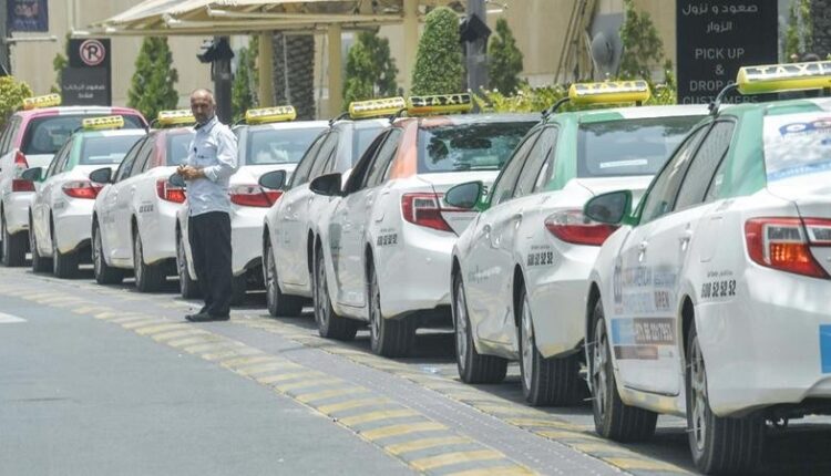 A row of official white Sharjah taxis lined up at a designated passenger pickup lane with a driver standing nearby.|Arabwheels.ae