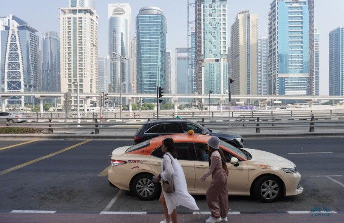 سيارة أجرة دبي بلون كريمي وبرتقالي مركونة على طول شارع في المدينة بينما تمر امرأتان بجانبها على الرصيف. وفي الخلفية، يرتفع أفق كثيف من ناطحات السحاب الحديثة خلف جسر علوي لطريق سريع متعدد المسارات تحت سماء صافية. | Pakwheels.com
