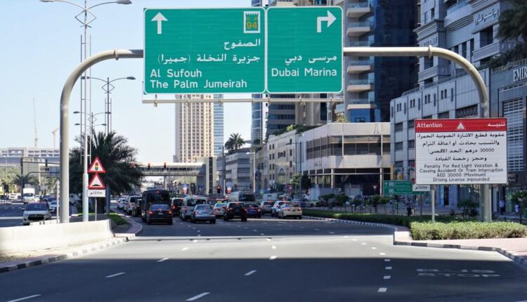 Busy urban road with multiple lanes, overhead directional signs, traffic, and a roadside warning notice.