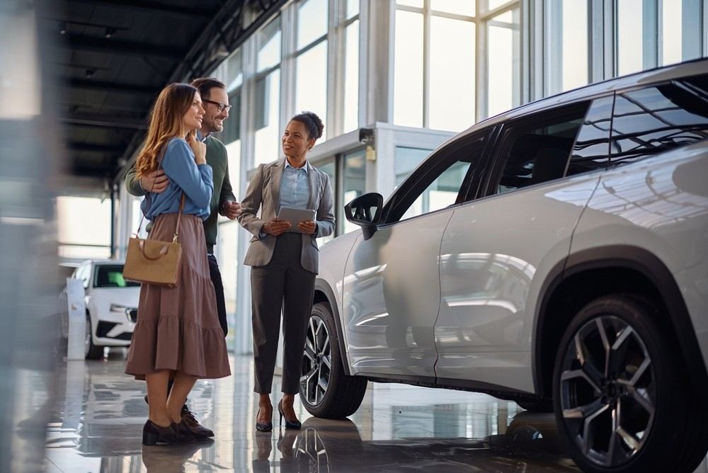 Couple speaking with a salesperson beside a new SUV in a showroom during the UAE new-car price war.|Arabwheels.ae